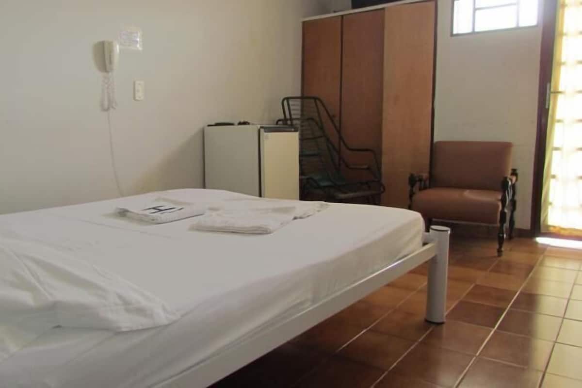 Une chambre avec un lit simple, recouverte d’une courtepointe blanche, une petite table de chevet, une chaise et une armoire en arrière-plan.