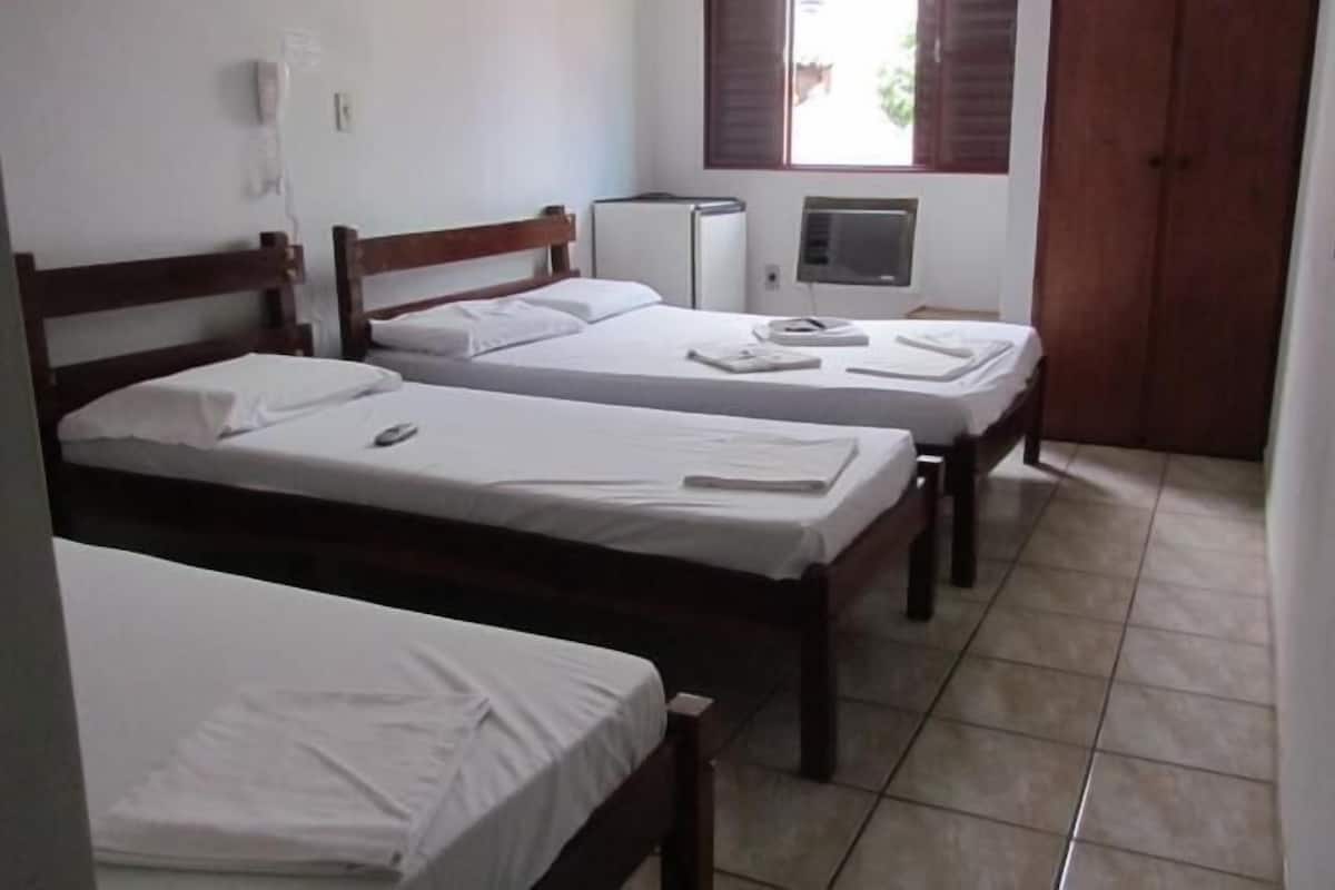 Une chambre avec trois lits simples, un petit réfrigérateur blanc, un radiateur et une fenêtre avec des stores.