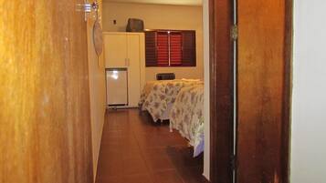A narrow hallway with a wooden door on the left, a small refrigerator, and a bed with floral bedding in the background.