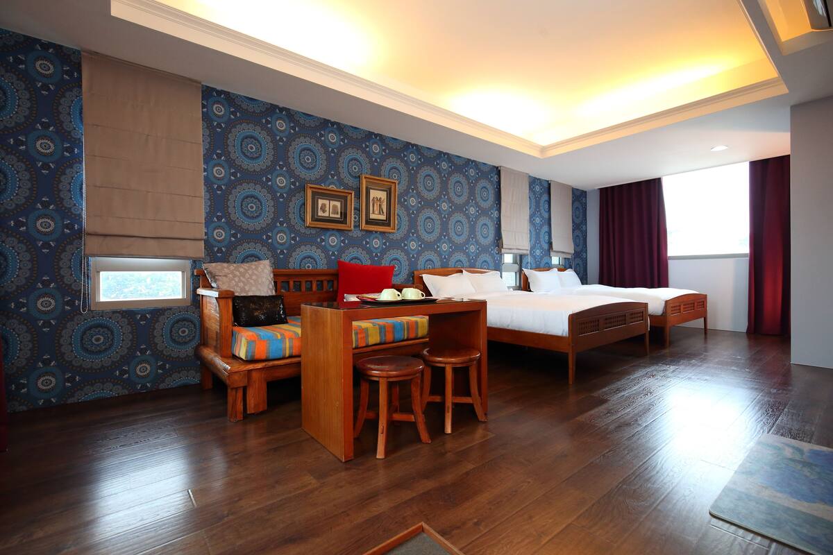 A hotel room with a bed, a desk with a chair, a small table, and a window with curtains.