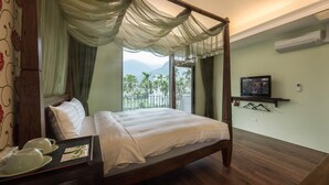 Una cama con dosel y cortinas traslúcidas, un televisor en un estante y un balcón con vistas a palmeras y montañas.