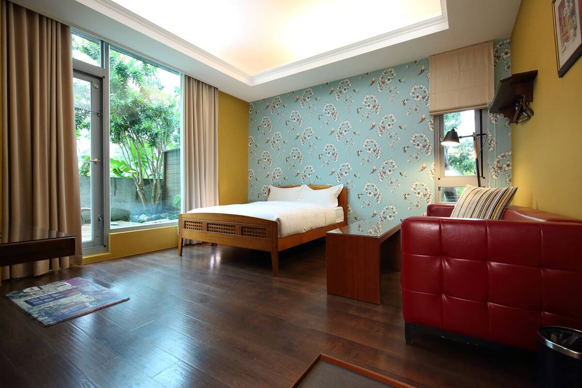 A bedroom with a bed, a red sofa, a window with curtains, and a wooden floor.