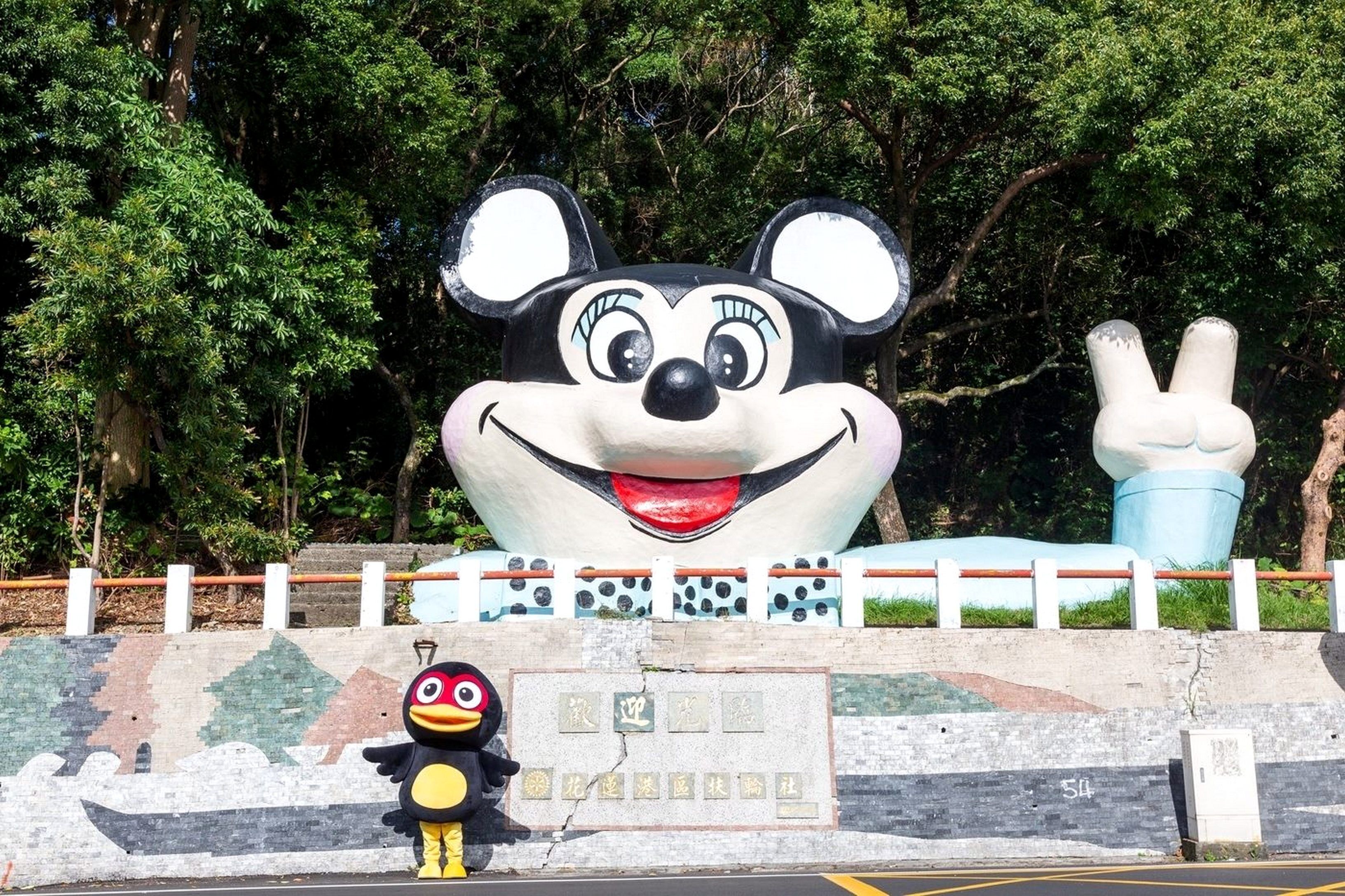 A large Mickey Mouse statue with a peace sign, a bird mascot, and a plaque with Chinese characters.