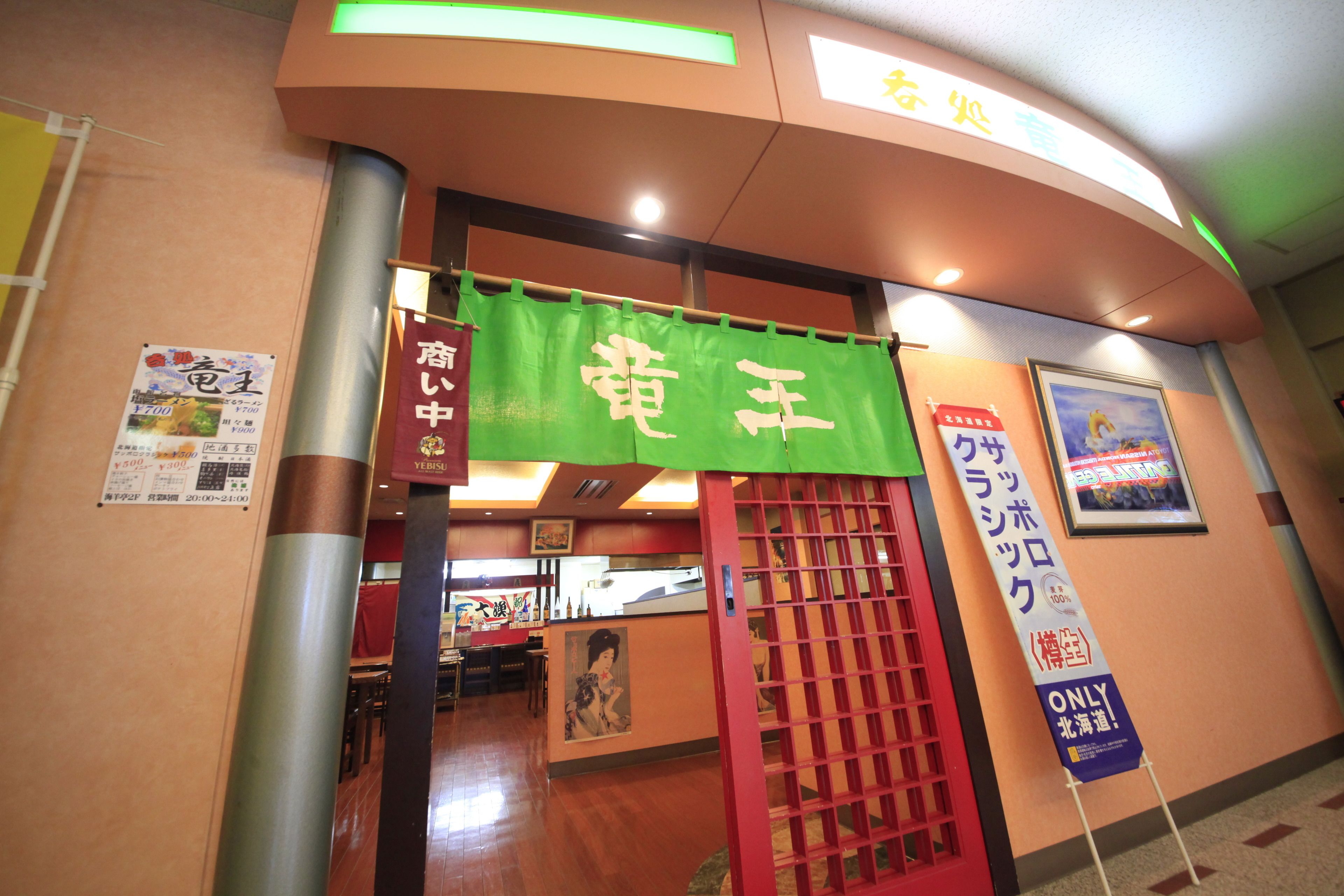 Entrada a un restaurante con toldo verde mostrando caracteres japoneses, una puerta roja y un cartel en la pared.