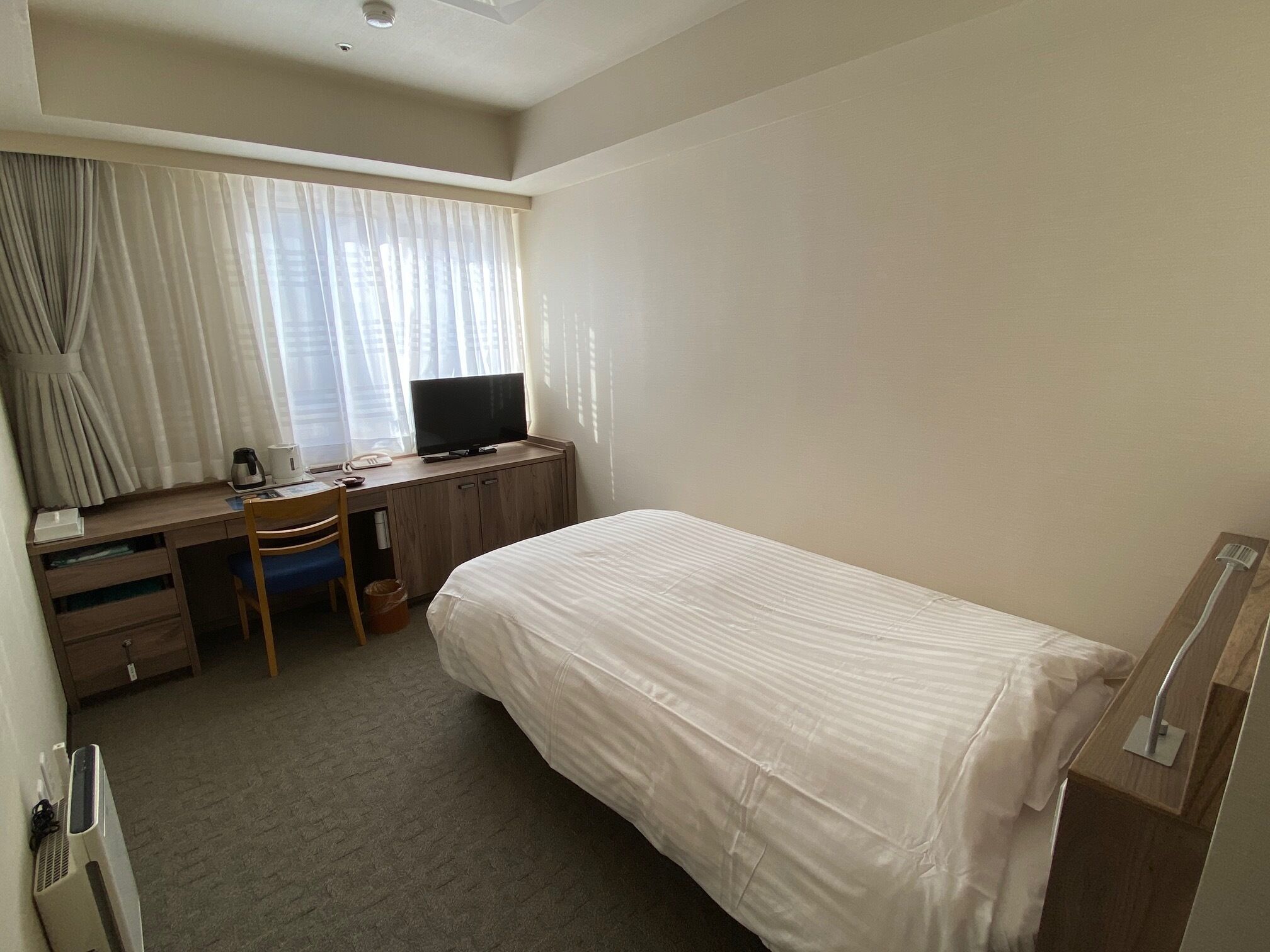 A hotel room with a bed, a desk with a computer, a chair, a window with curtains, and a bedside table.