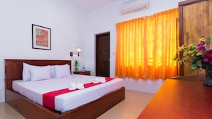 Una habitación de hotel con una cama de madera, una colcha roja y blanca, una cortina naranja, una mesita de noche de madera con una lámpara, y un jarrón con flores.