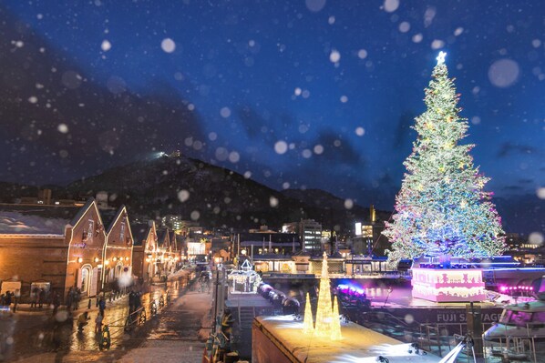 Rua coberta de neve com uma grande árvore de Natal iluminada e edifícios tradicionais.
