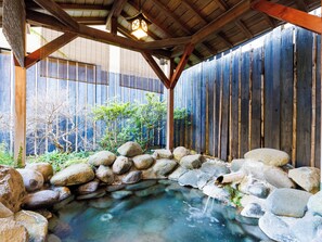 Uma onsen japonesa tradicional com um deck de madeira, uma banheira de pedra rodeada por pedras e uma cerca de bambu ao fundo.