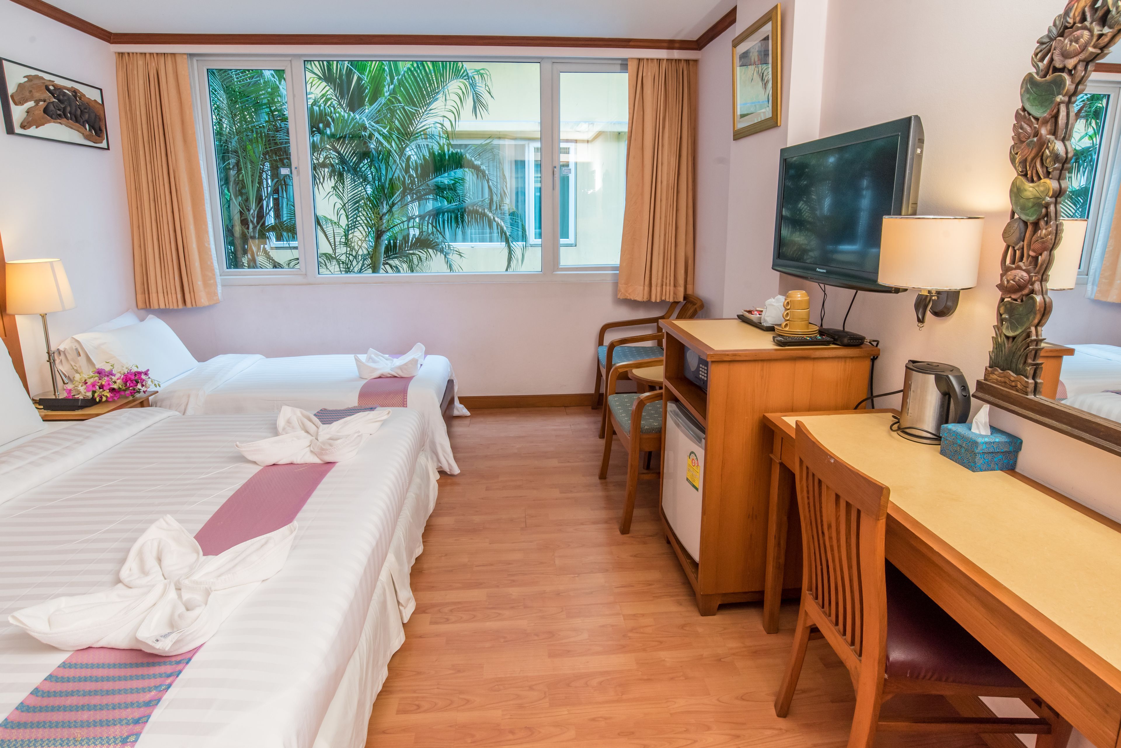 A hotel room with two beds, a desk, a television, and a window with a view of palm trees.