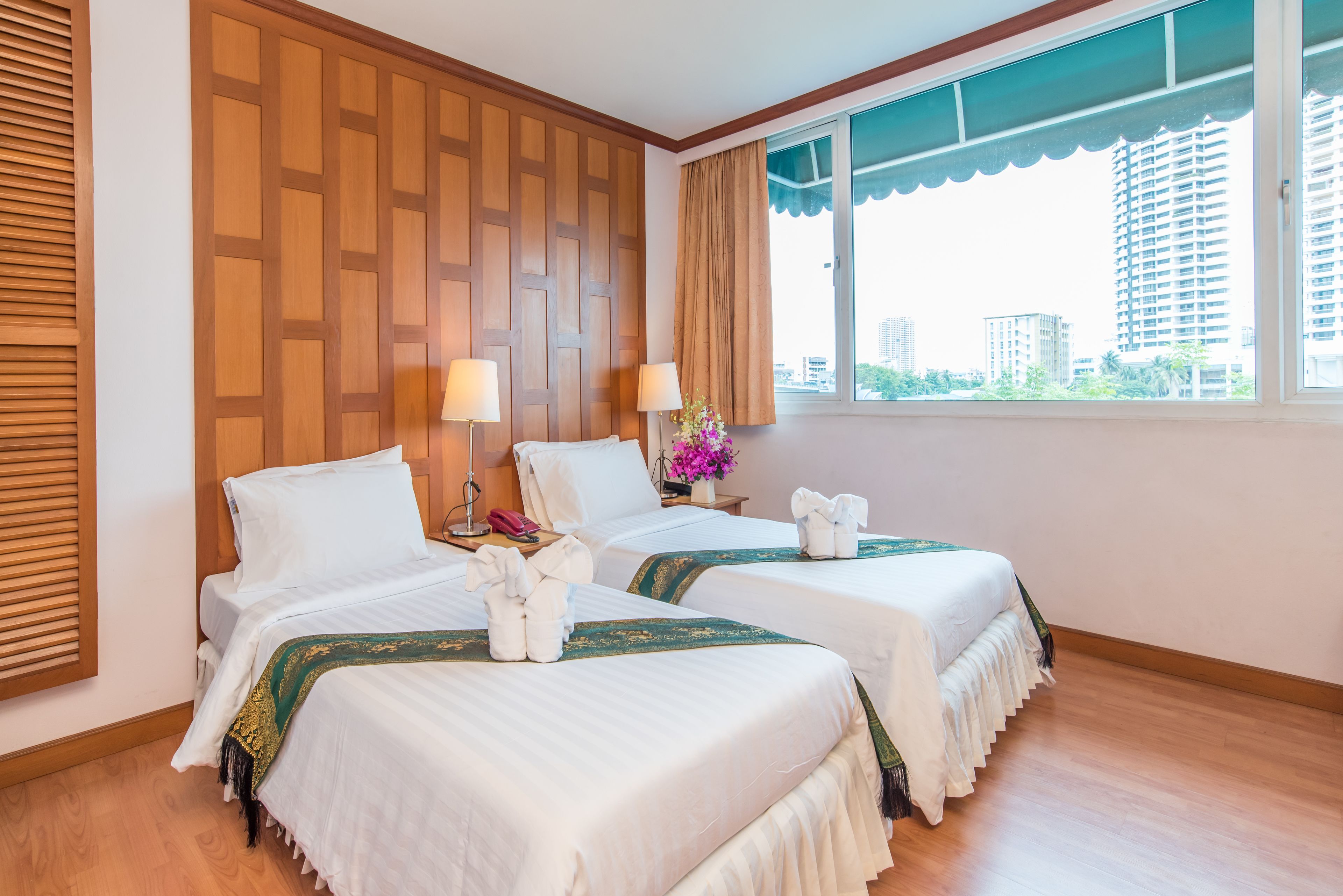 A hotel room with two beds, wooden paneling on the wall, a window with a view of buildings, and a flower vase on the nightstand.