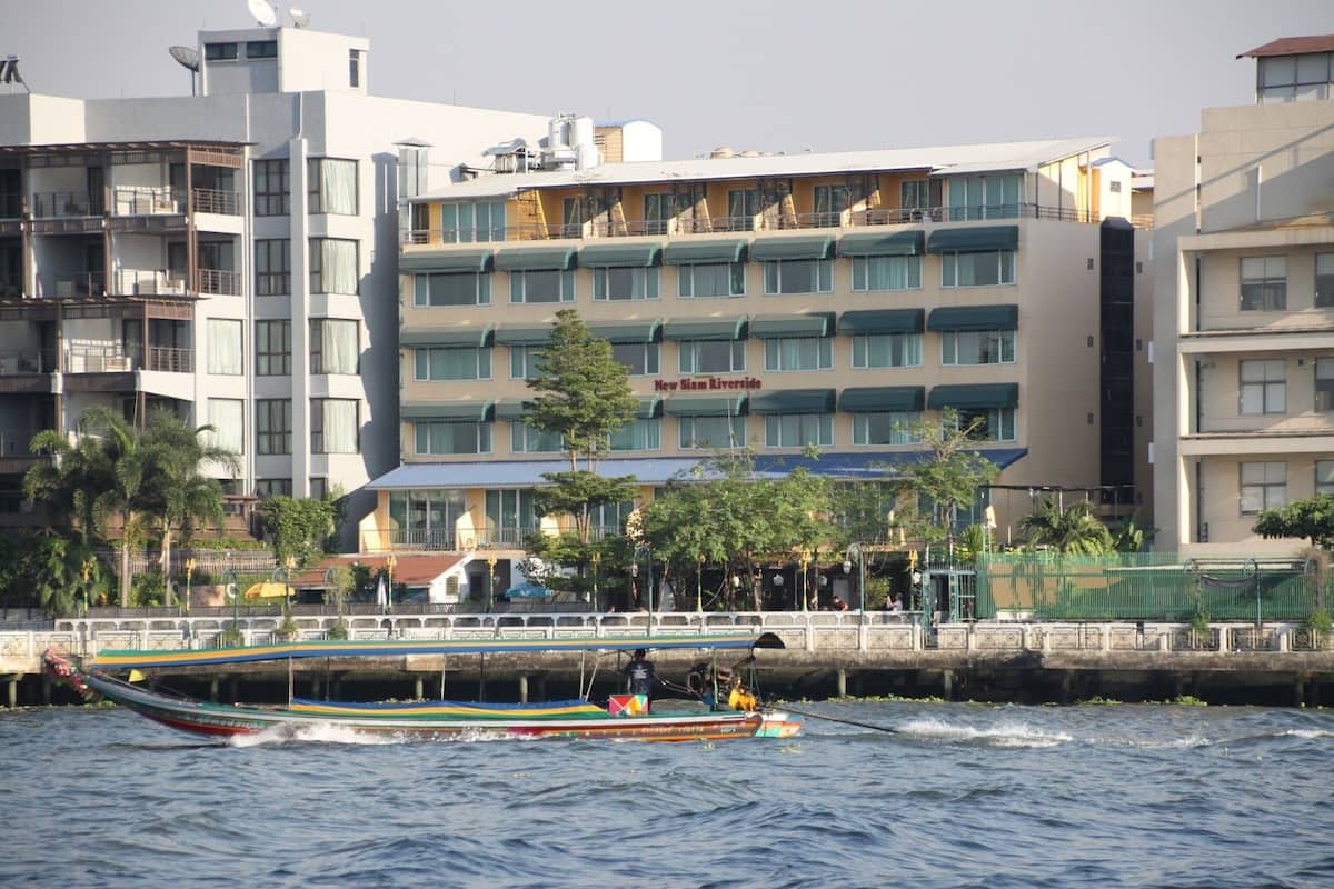 A boat with passengers, a building with balconies, and a sign reading "New Siam Riverside".