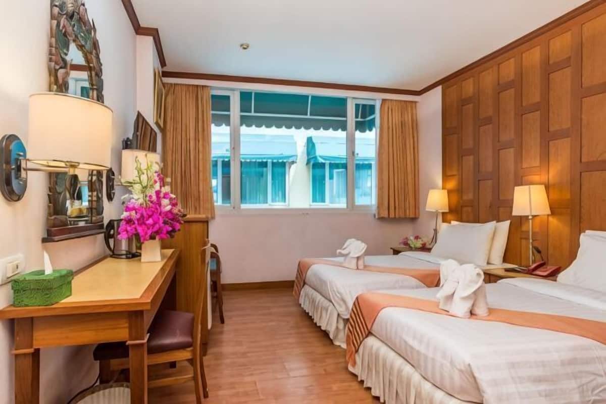 A hotel room with two beds, a desk with a lamp, a chair, a window with curtains, and a wooden wall paneling.