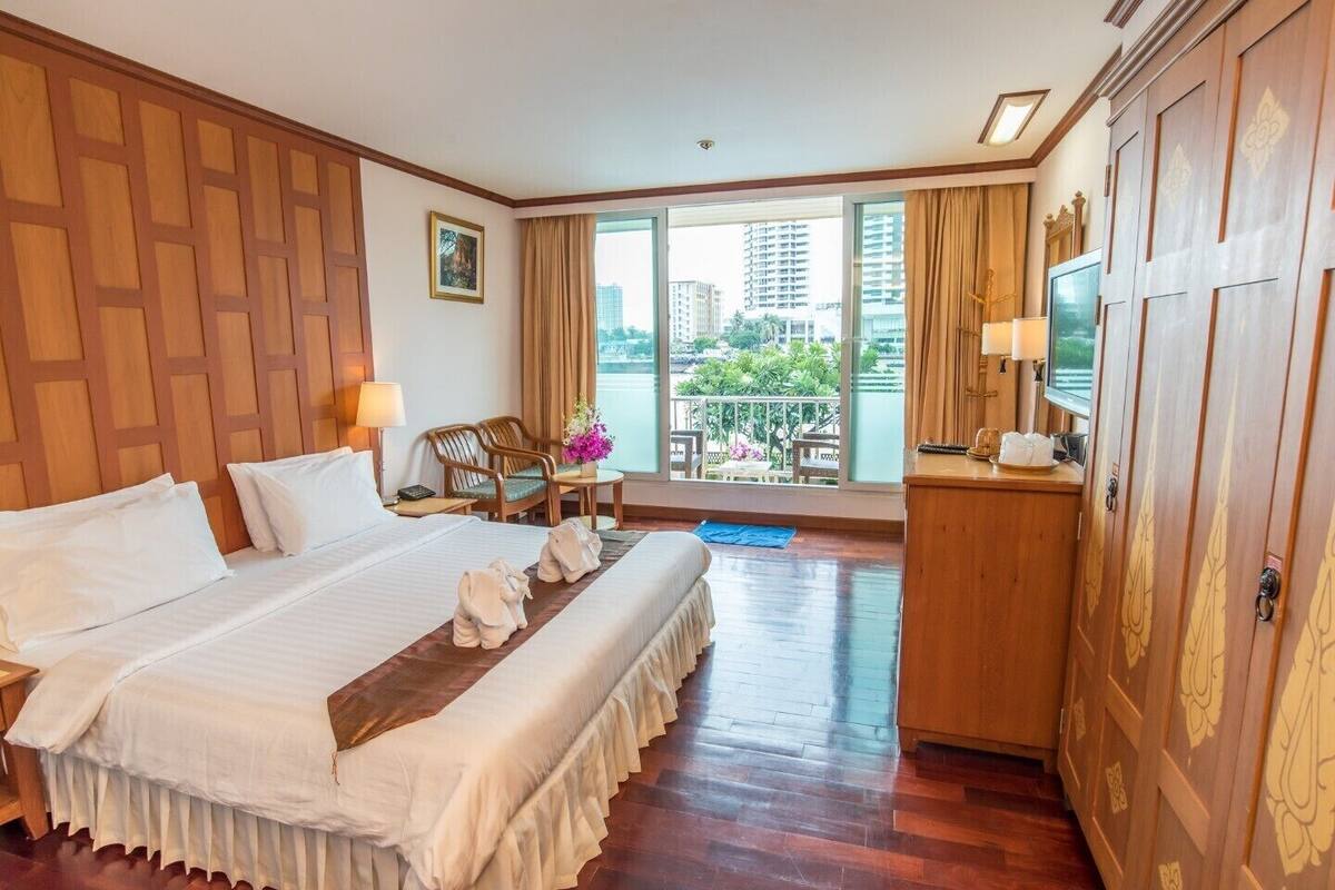 A hotel room with a large bed, wooden paneling, a desk with a TV, and a balcony with a view of buildings.