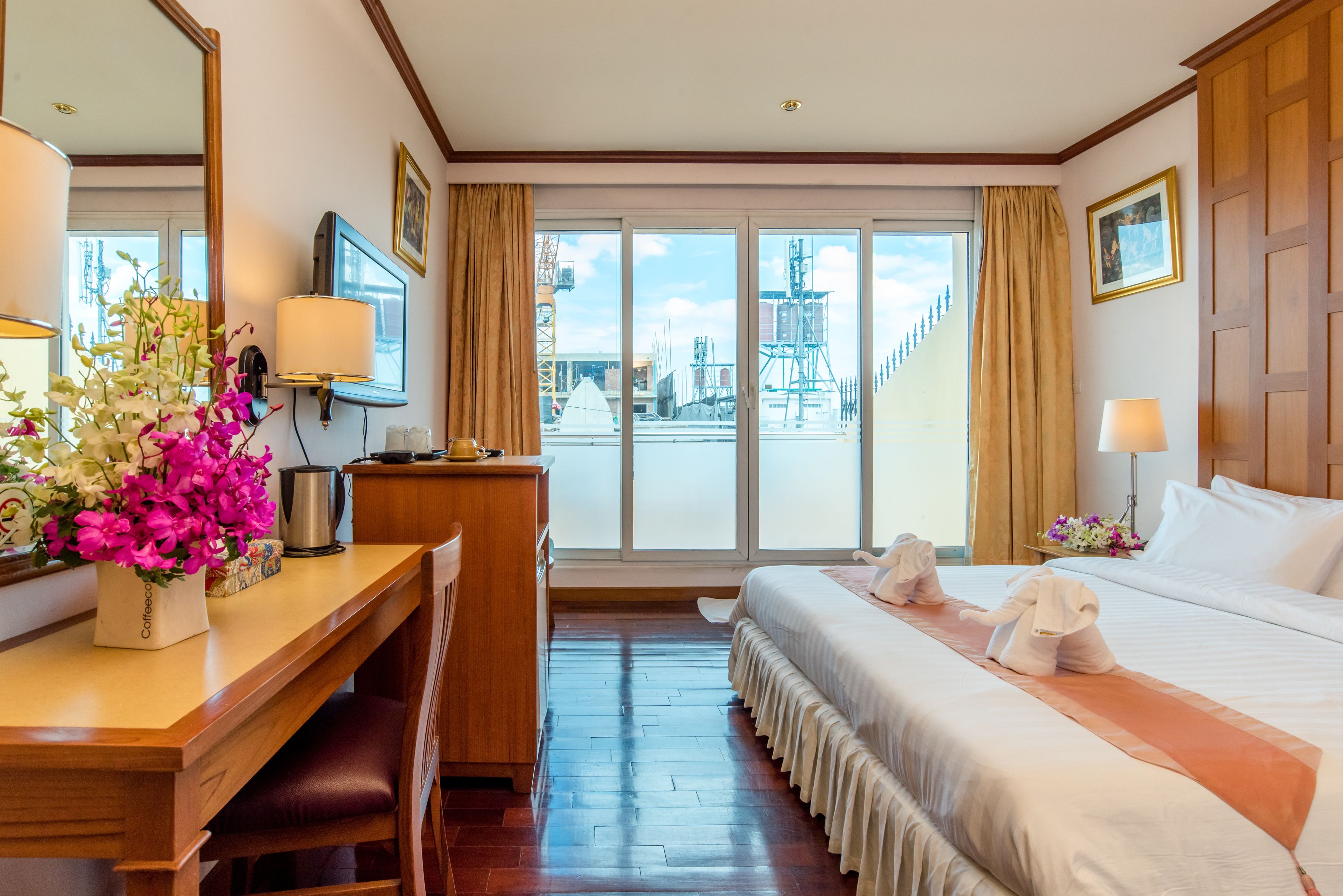 A hotel room with a bed, a desk, a chair, a coffee maker, a vase with flowers, a lamp, and a large window with a view of a building.