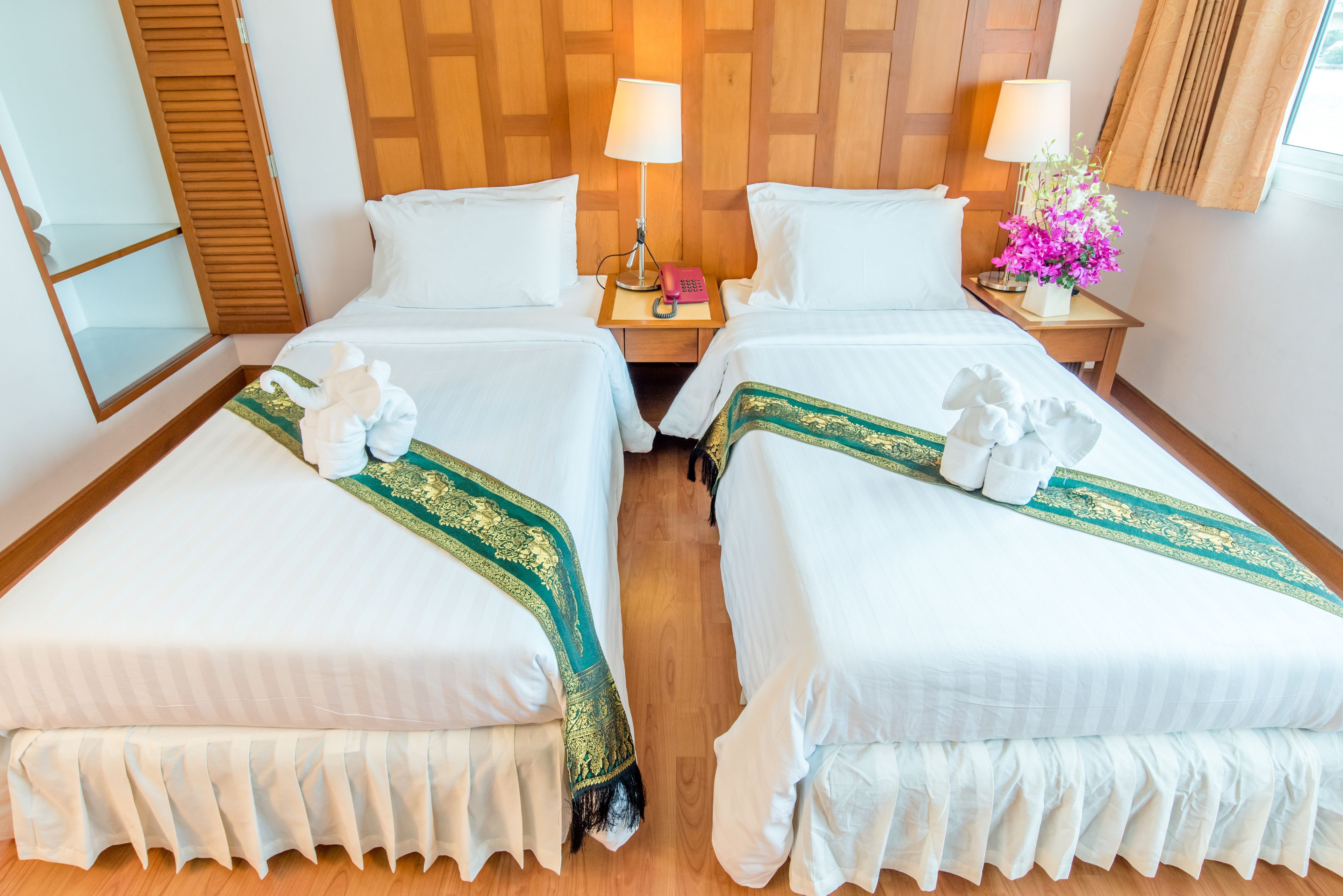 A hotel room with two beds, each with white bedding and decorative pillows. There are bedside tables with lamps and a small table with a pink telephone and a vase of flowers.
