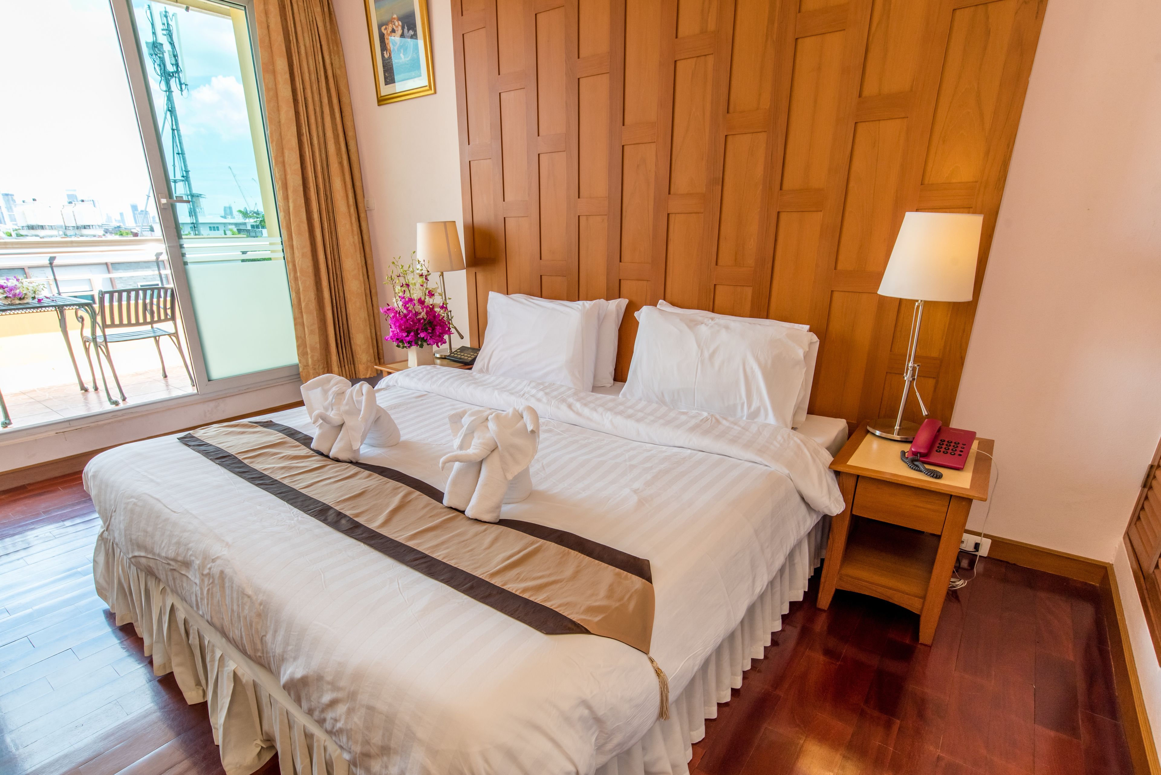 A hotel room with a large bed, wooden headboard, two bedside tables with lamps, a small table with a phone and a pink item, and a balcony with a view of the city.