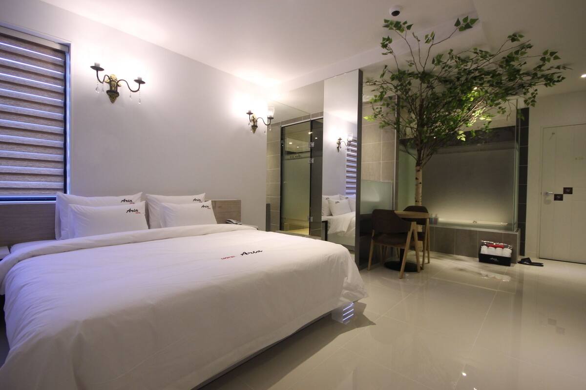 A modern hotel room with a large bed, a small desk, a chair, a potted plant, and a glass-enclosed shower area.