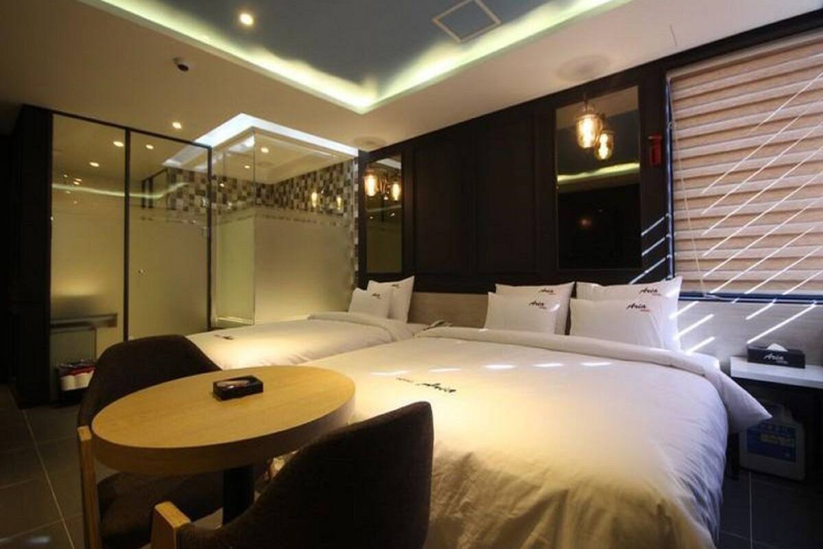 A modern hotel room with a large bed, a round table, and a chair. There is a glass partition and a window with blinds.