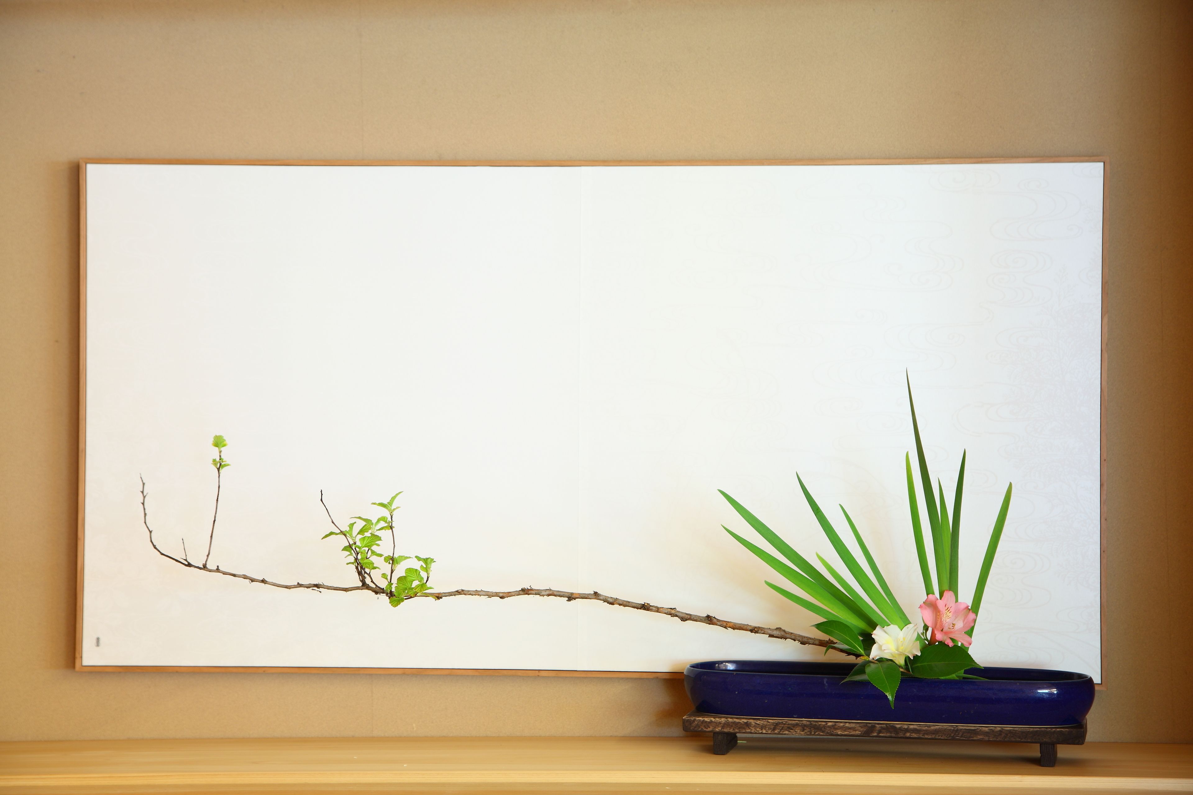 A branch with green leaves and pink flowers in a blue vase on a shelf.