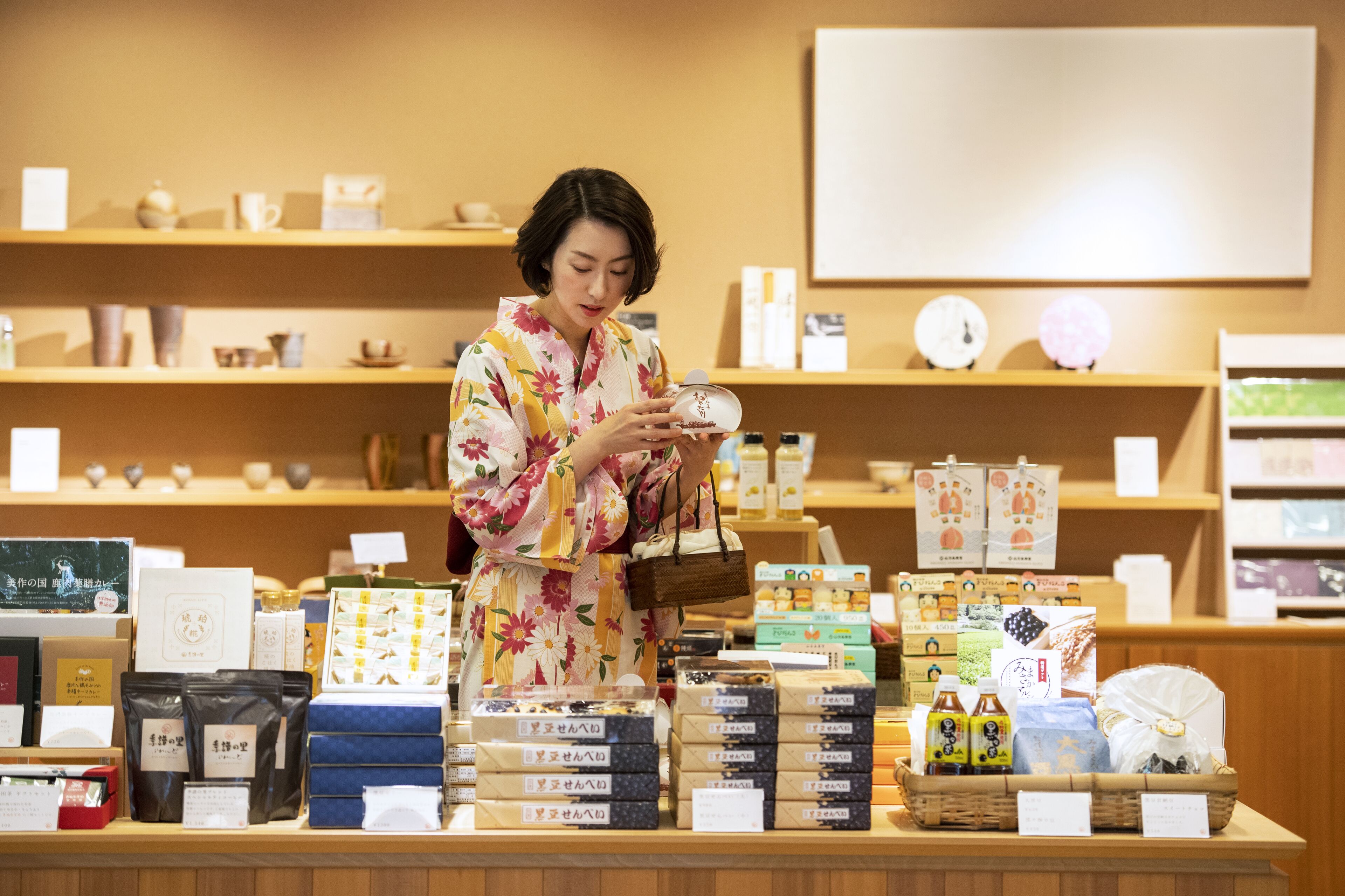 A woman in a floral kimono examining a product on a display counter in a store with various packaged goods and items on shelves.