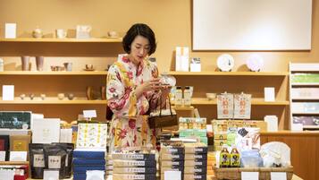 A woman in a floral kimono examining a product on a display counter in a store with various packaged goods and items on shelves.