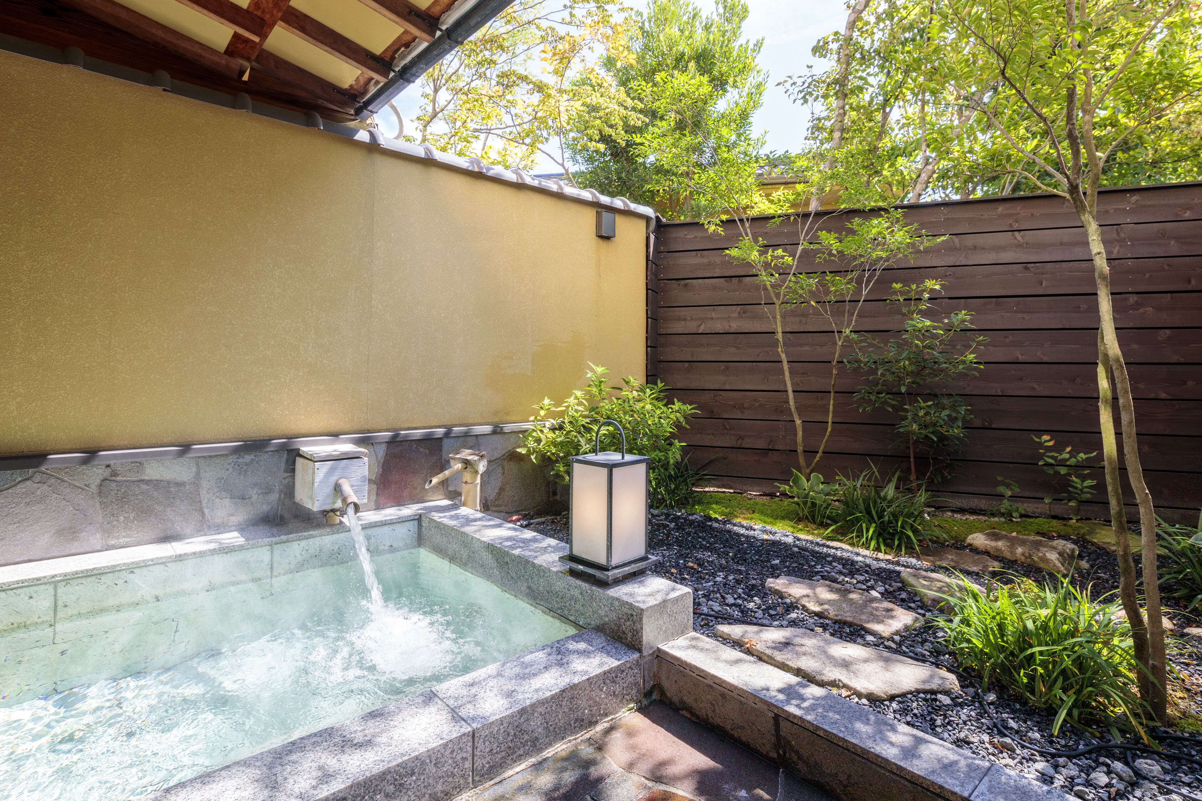 Un jacuzzi con borde de piedra, cerca de madera y un pequeño jardín con plantas y piedras.