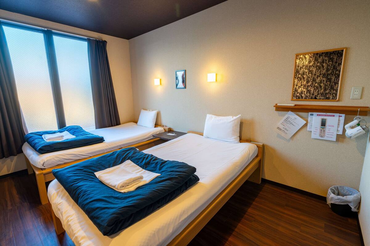 Ett hotellrum med två separata sängar, ett stort fönster med gardiner, väggmonterade lampor, en inramad tavla och en liten hylla med papper.