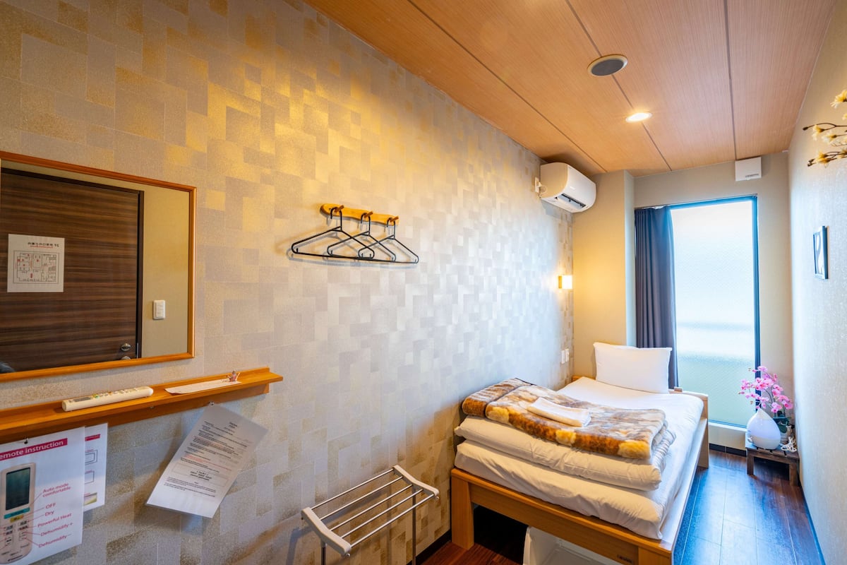 Ett hotellrum med en säng, en sänggavel i trä, en väggmonterad luftkonditioneringsanläggning, ett fönster med gardiner, en spegel och en hylla med en fjärrkontroll och ett papper.