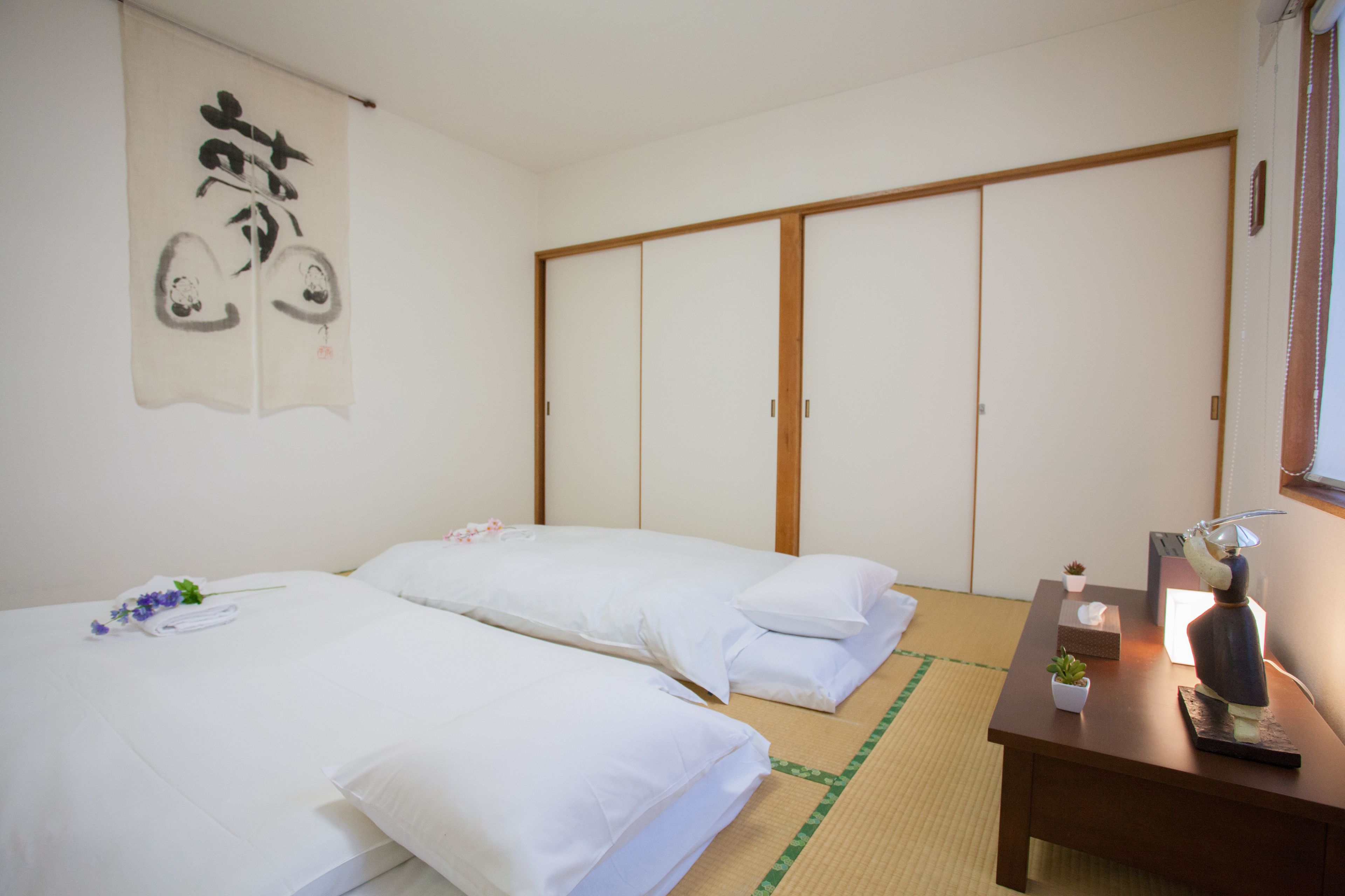 Una habitación japonesa tradicional con suelo de tatami, dos camas individuales, un armario de madera y una mesita con un florero.