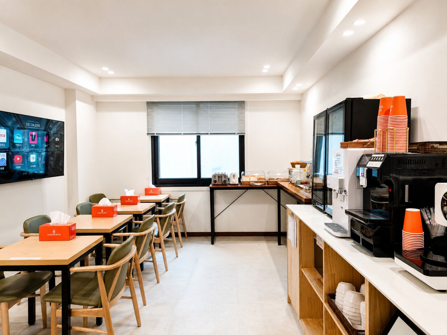 A modern hotel room with a flat-screen TV, a coffee machine, and a dining area with tables and chairs.