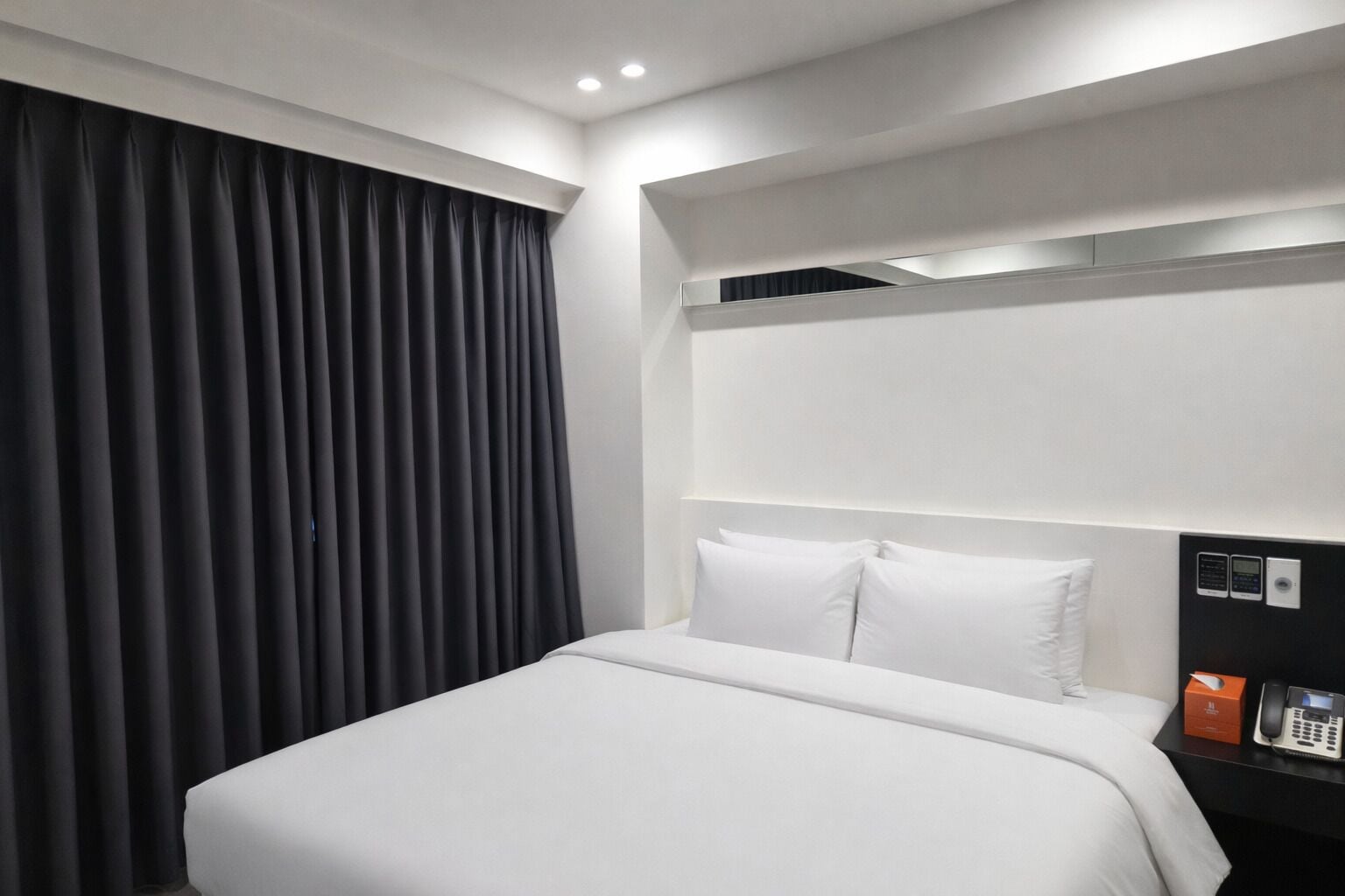 A modern hotel room with a large bed, a dark curtain, a bedside table with a phone, and a control panel.