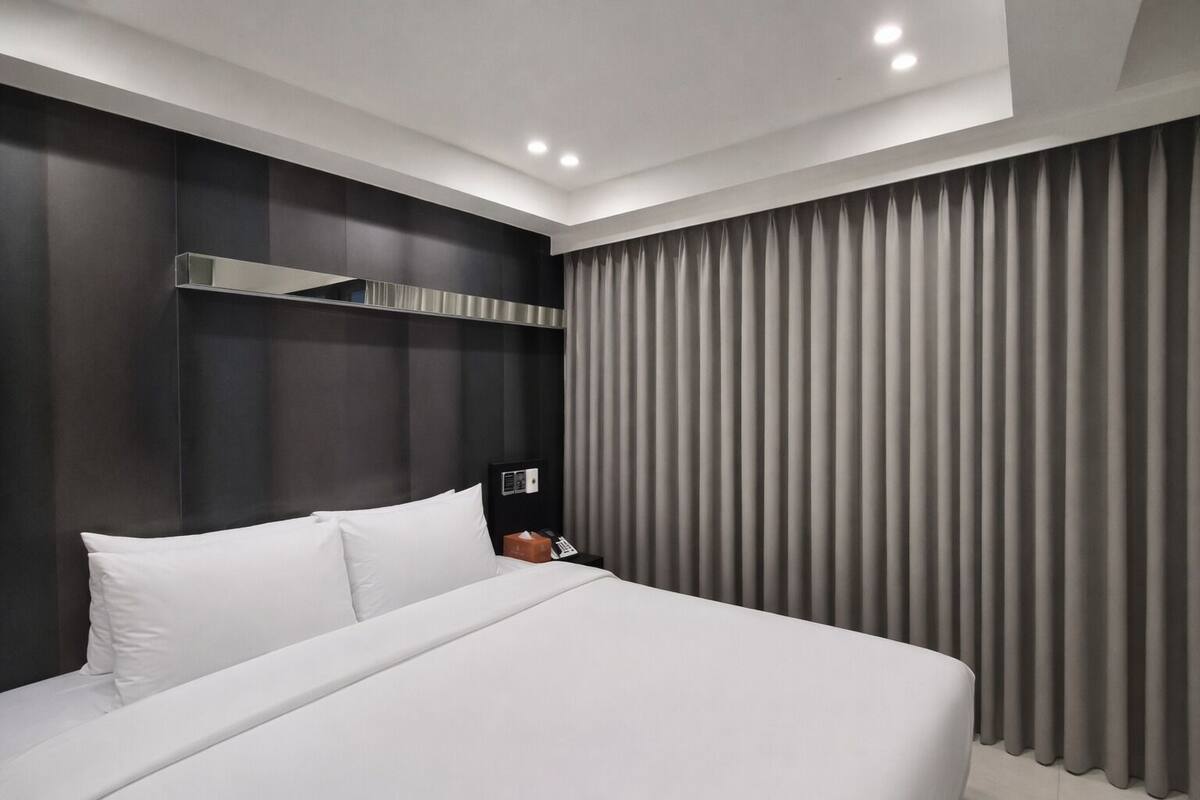 A modern hotel room with a large bed, dark wood paneling, a bedside table with a phone, and a window with vertical blinds.