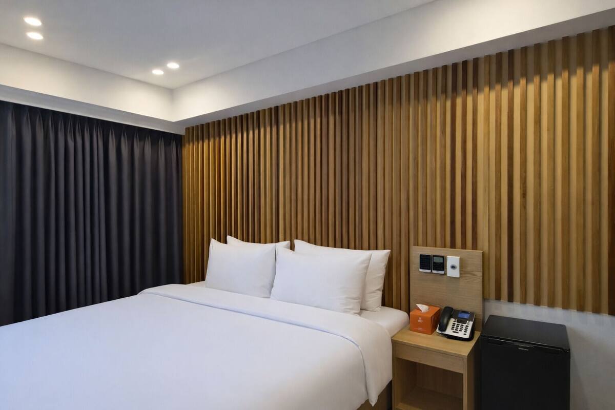 A hotel room with a bed, a wooden headboard, a bedside table with a phone and a small appliance, and a dark curtain.
