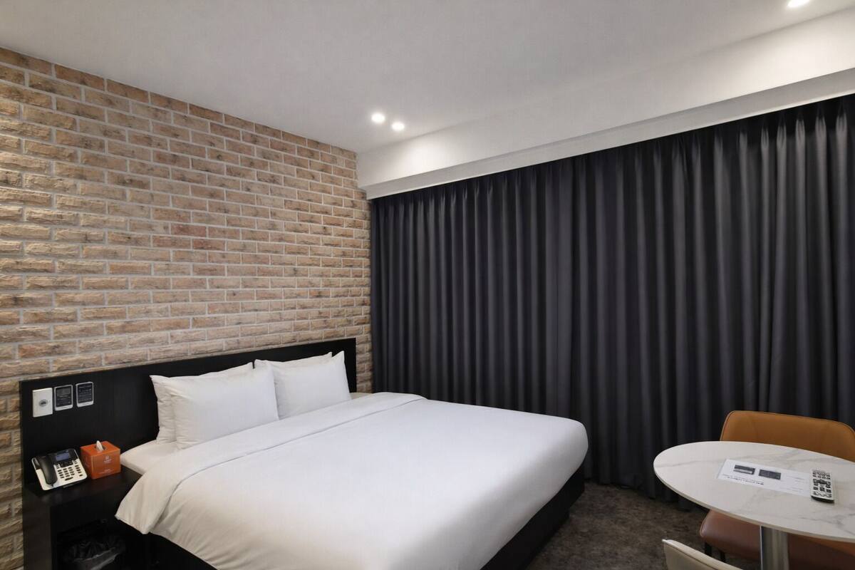 A hotel room with a brick wall, a bed with white linens, a black headboard, a small table with a chair, and a black curtain.