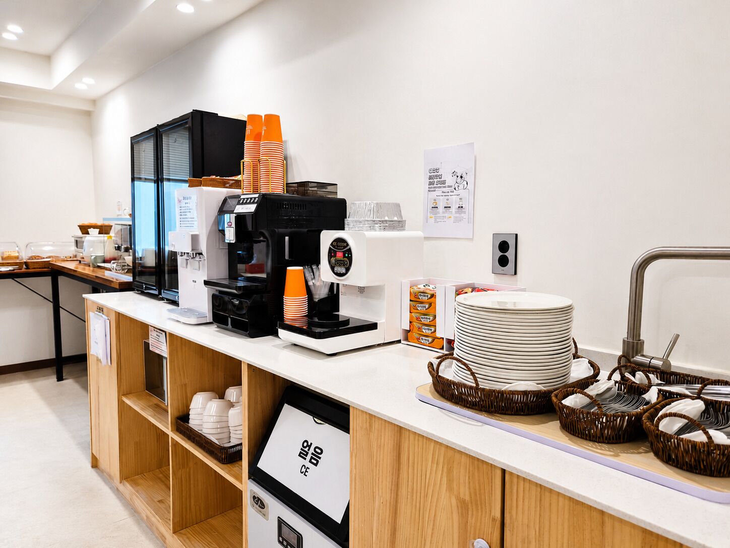 Una moderna area caffè con diverse macchinette, tazze arancioni e una pila di piatti bianchi.
