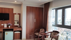 Quarto compacto com escrivaninha de madeira, televisão, pequena geladeira e área de refeições com duas cadeiras.