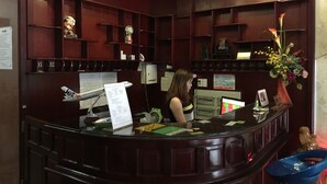 A reception desk with a computer, a telephone, and a menu board. There are shelves with various items and a statue on the right. - Huong Mai 2 Hotel (Ho Chi Minh City)