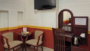 A hotel room with a brick-patterned wall, a flat-screen TV mounted above a wooden table with two chairs, a mirror, and a small shelf with a kettle and a sign. - Huong Mai 2 Hotel (Ho Chi Minh City)