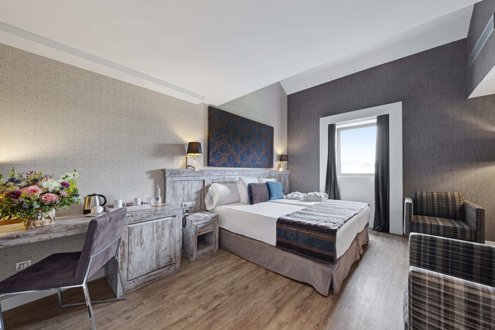 Habitación de hotel moderna con una cama grande, un escritorio con una silla, una mesita con un jarrón de flores y una ventana con vistas.