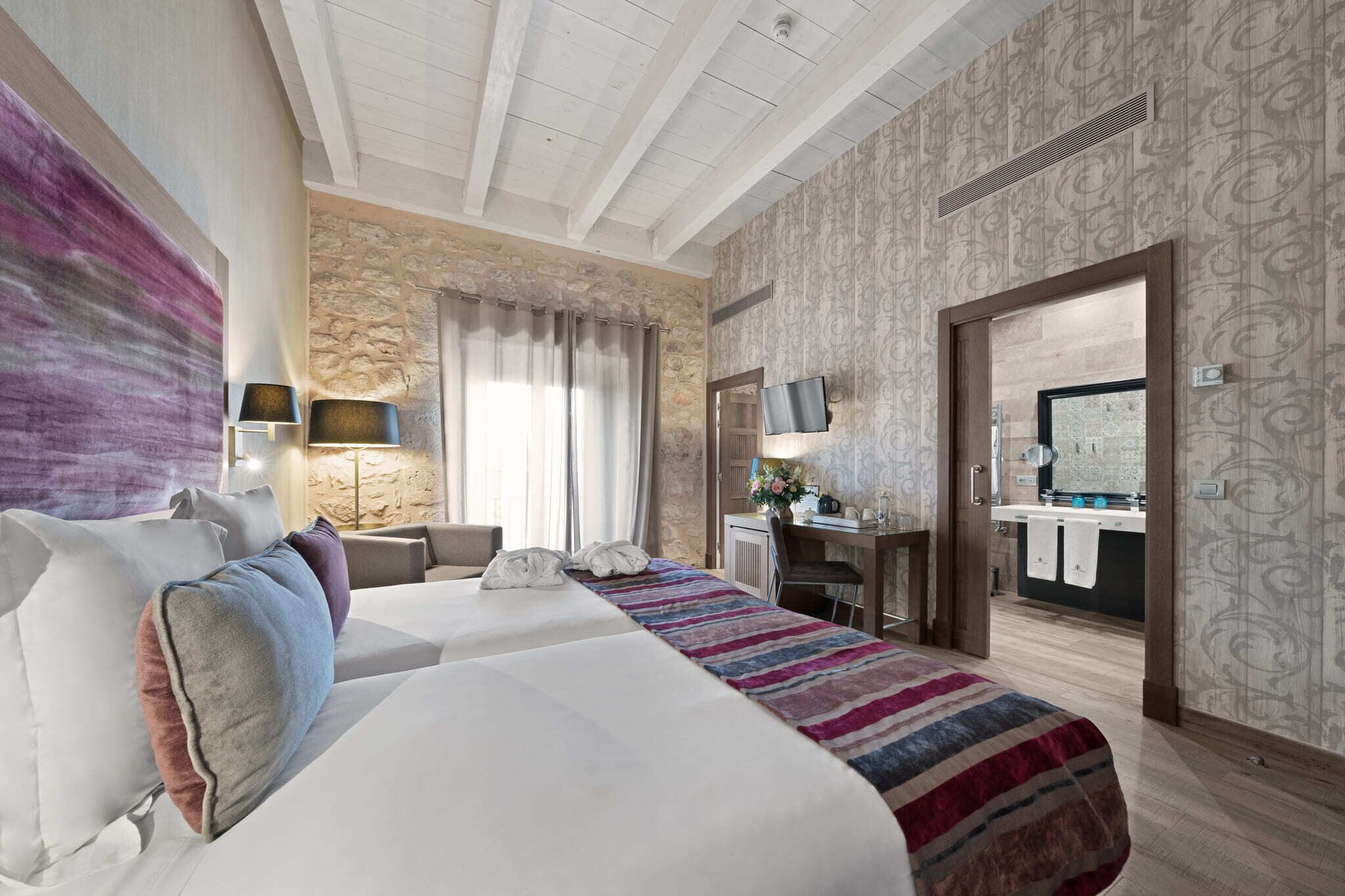 A hotel room with a large bed, a desk, a TV, and a stone wall.