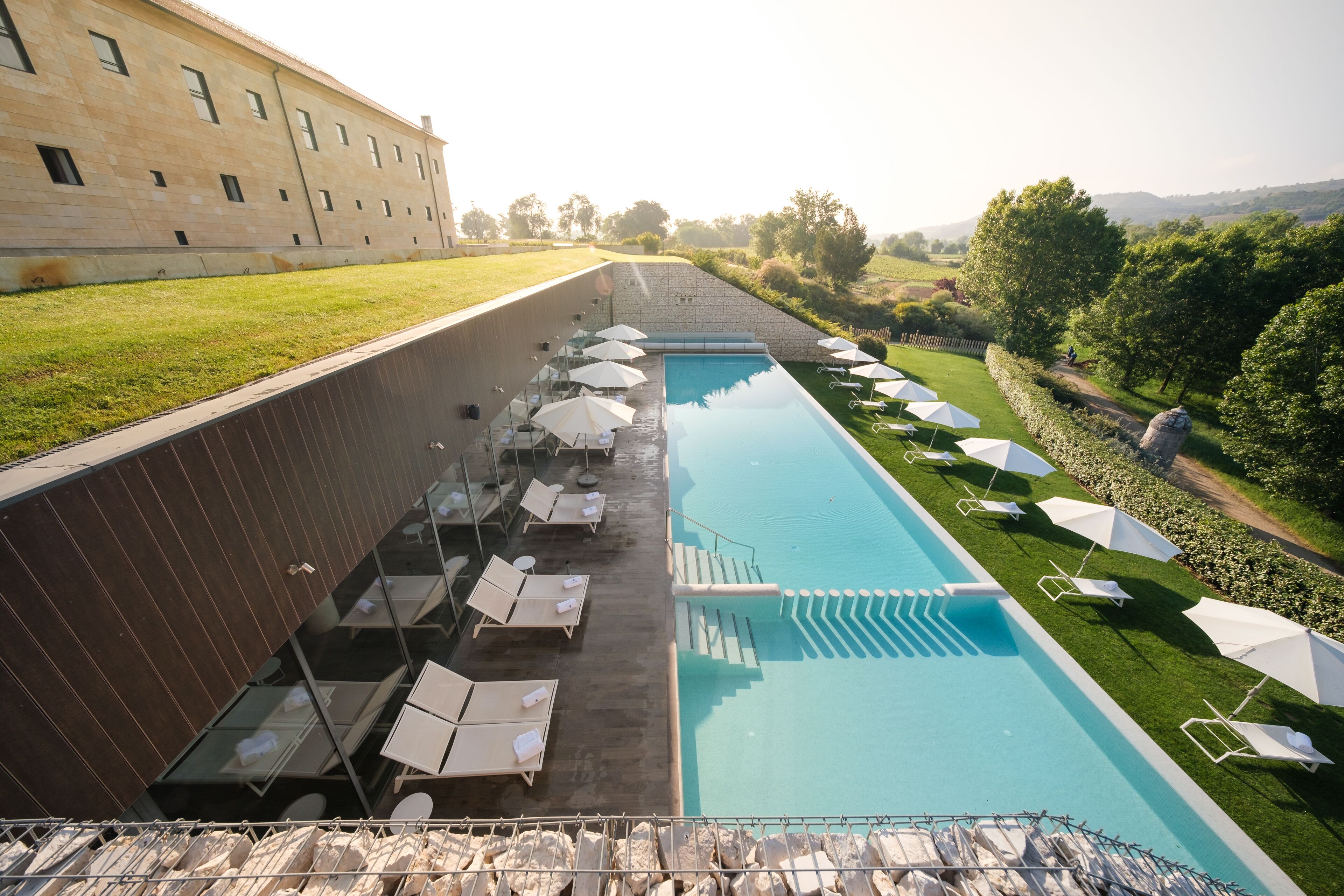 Ein Swimmingpool mit Liegestühlen und Sonnenschirmen, umgeben von einer Steinmauer und einer Rasenfläche.