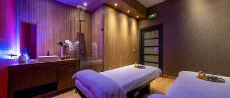 Un cuarto de spa con dos camillas de masaje, paneles de madera, un tocador con espejo y un pequeño rincón de descanso.