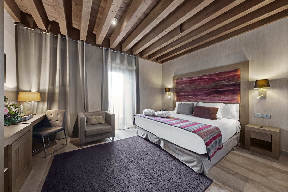 A modern hotel room with a wooden ceiling, a large bed with a colorful headboard, a desk with a lamp, and a chair.