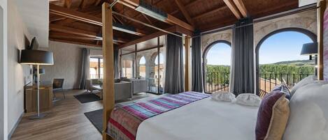 A spacious bedroom with a large bed, wooden beams, a balcony with a view, and a desk area.