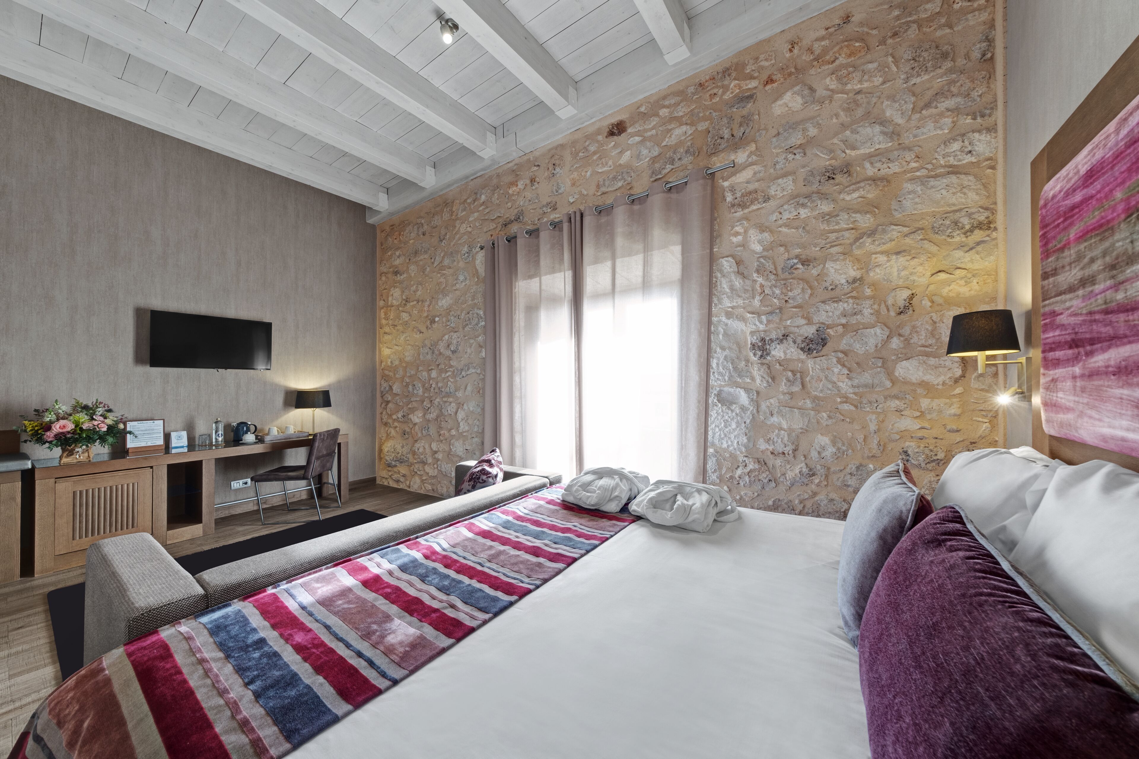 A hotel room with a stone wall, a bed with a striped blanket, a desk with a computer, and a TV mounted on the wall.