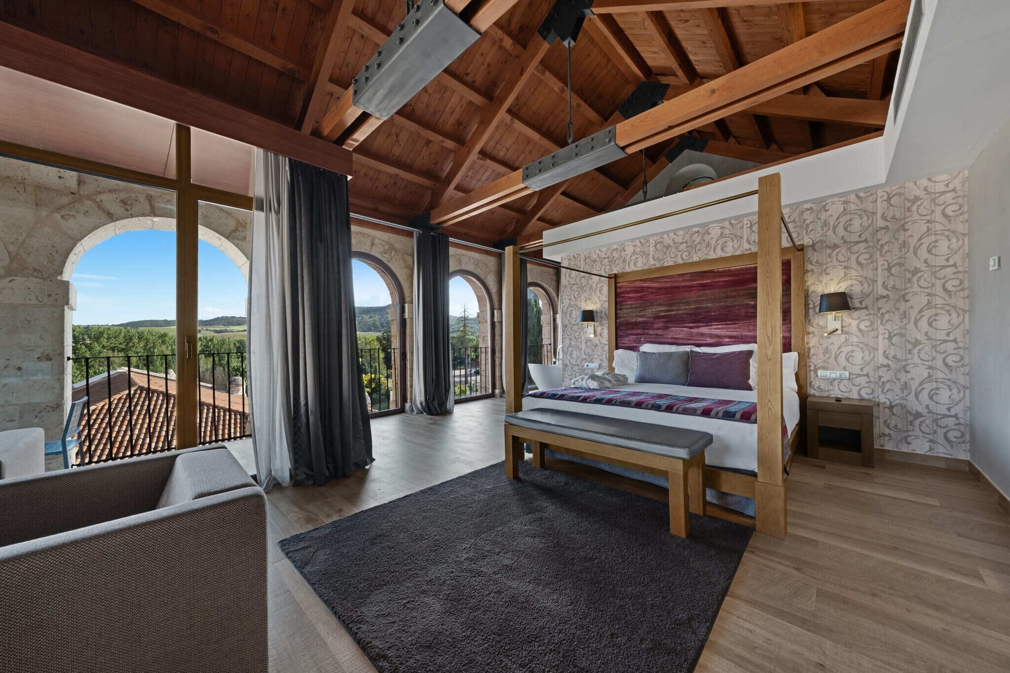 A bedroom with a large bed, wooden beams, a balcony, and a view of the countryside.