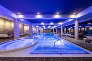Una amplia piscina interior con jacuzzi, rodeada de sillones de descanso y un muro de piedra.