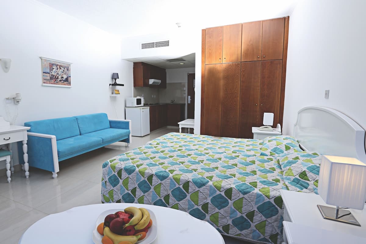Une chambre d’hôtel comprenant un lit, un canapé bleu, une petite table avec un panier de fruits, un coin cuisine et une armoire.