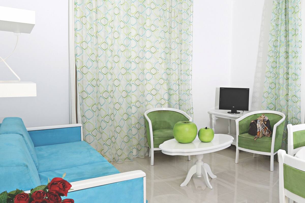 Une pièce moderne avec un canapé bleu, deux fauteuils verts, une petite table blanche sur laquelle se trouve un ordinateur portable, et un rideau à motifs.