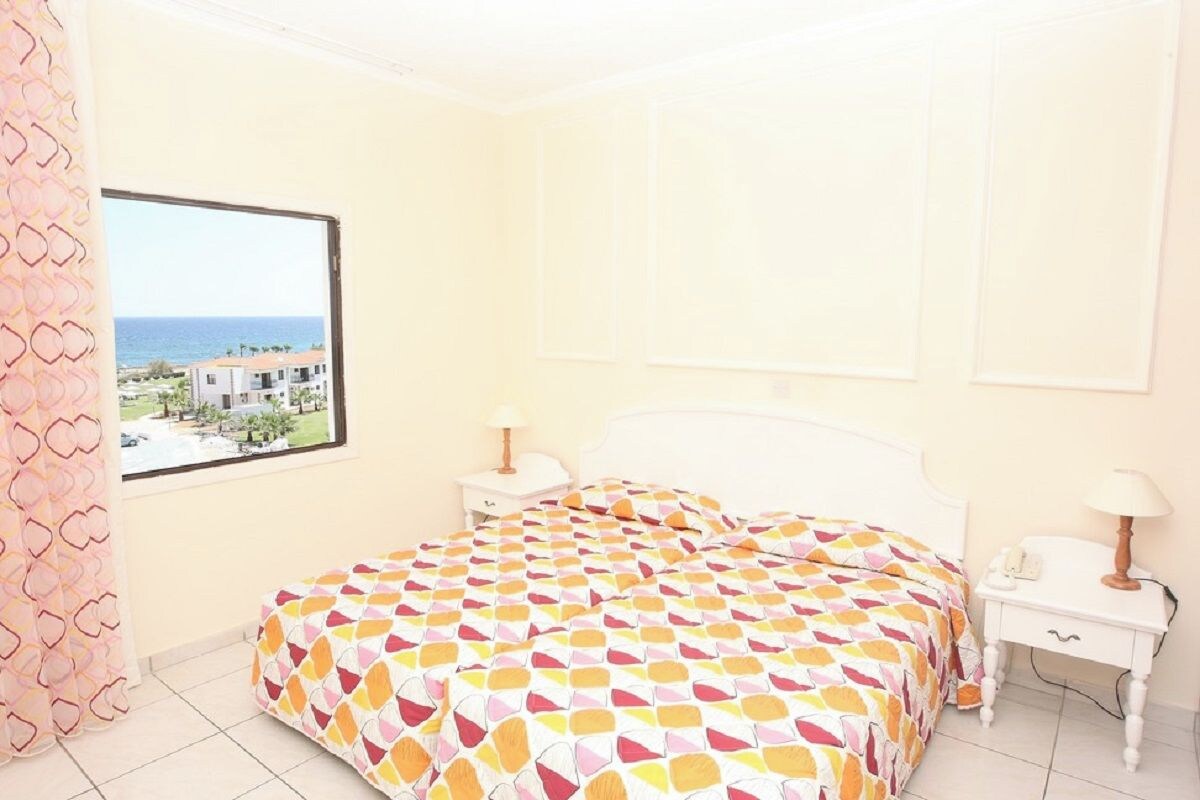 A bedroom with a bed, a nightstand, a lamp, and a window with a view of the sea and buildings.